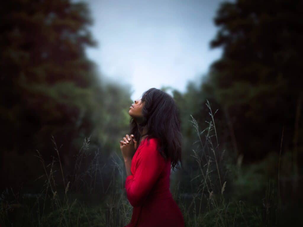 an image of a woman in red standing in a field with her eyes closed, looking up to the sky, and her hands clasped together in prayer