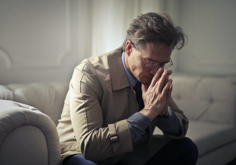 A mature man with glasses sits on a couch and clasps his hands to his face as if in prayer. He has a worried expression.