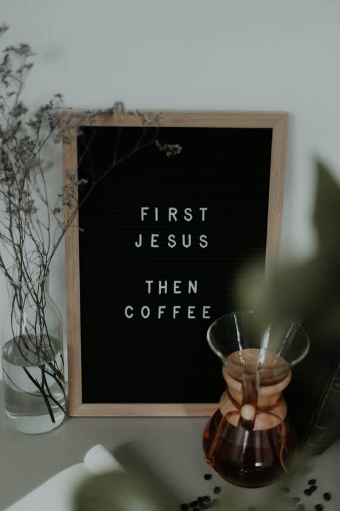 A felt letter board that reads "First Jesus, Then Coffee" placed behind a carafe of coffee.
