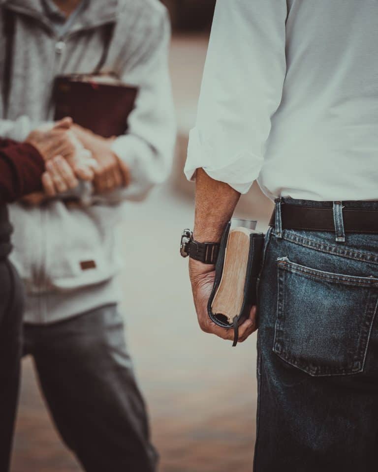 On the left stands two people. Their hands are pictured and one of them holds what appears to be a bible. On the right is a man in a white shirt and denim jeans holding a bible tucked to his side.