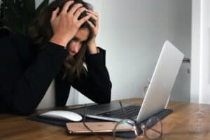 A woman sits in front of her laptop with her hands on her head as if frustrated.