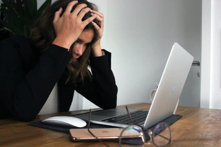 A woman sits in front of her laptop with her hands on her head as if frustrated.