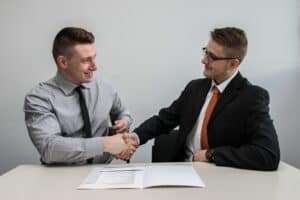 Two men sit at a table dressed in business professional clothing. They are shaking hands and a folder is opened before them.