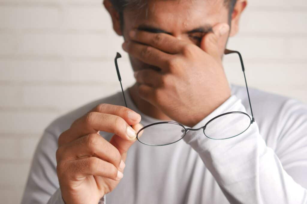 A man faces the camera. He hold his glasses in his right hand uses his left hand to pinch his eyes in frustration.