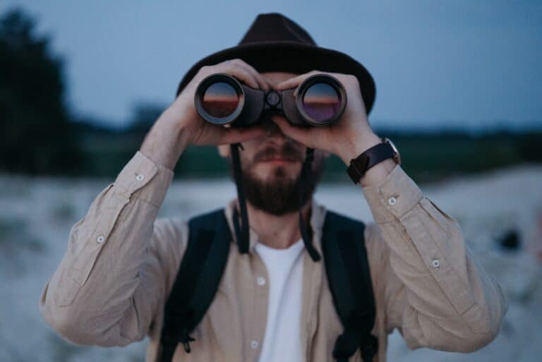 A man in a hat holds a pair of binoculars to his eyes while he looks at the camera