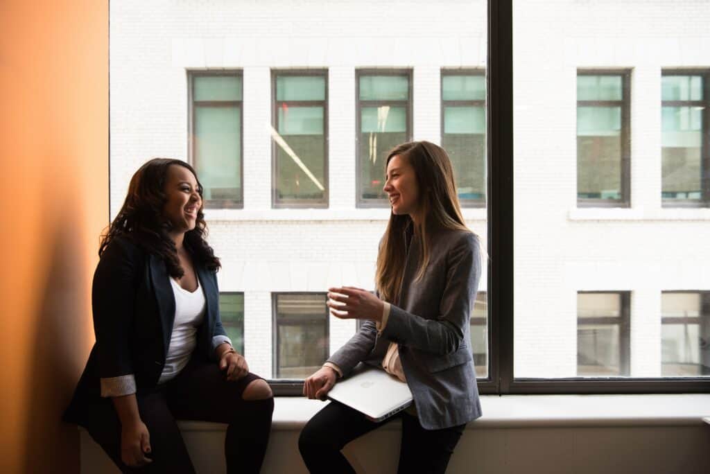 Two women sit on the ledge of an office window and are talking.
