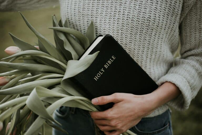 An image of a bible being held by someone. They also are holding flowers behind the bible.