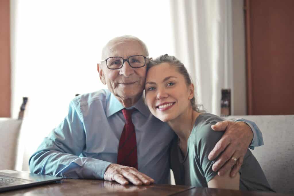 A mature man and younger woman share a hug while facing the camera