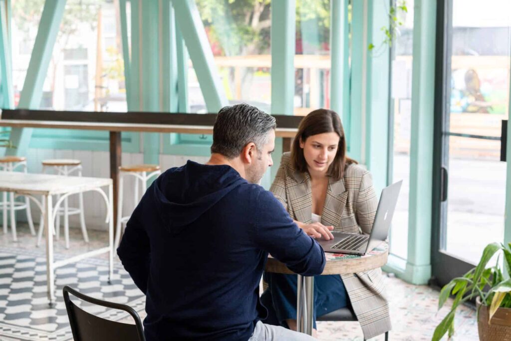 A man and woman sit at a table together looking at a laptop computer.