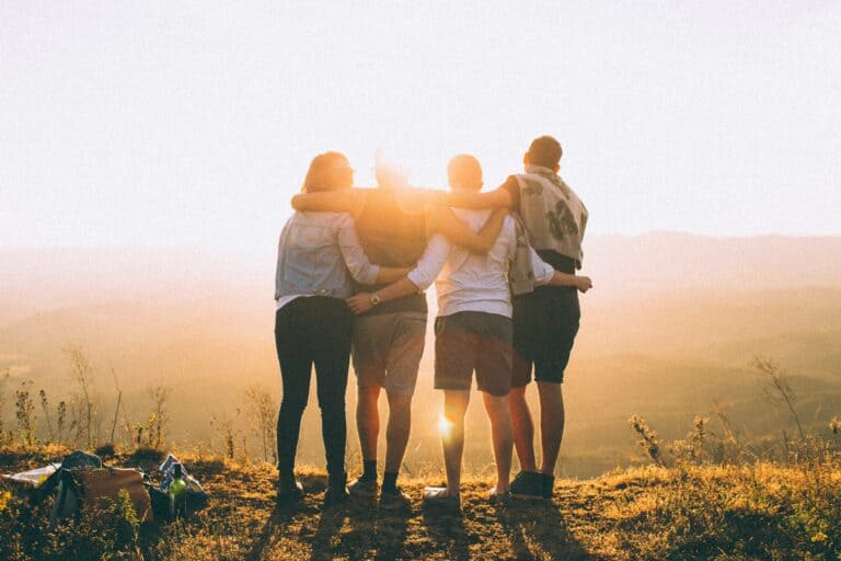 A group of people stand on top of a hill facing the sun with their arms around each other's necks.