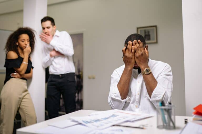 A man sits with his hands covering his face as a man and woman stand in the corner whispering and looking in his direction.