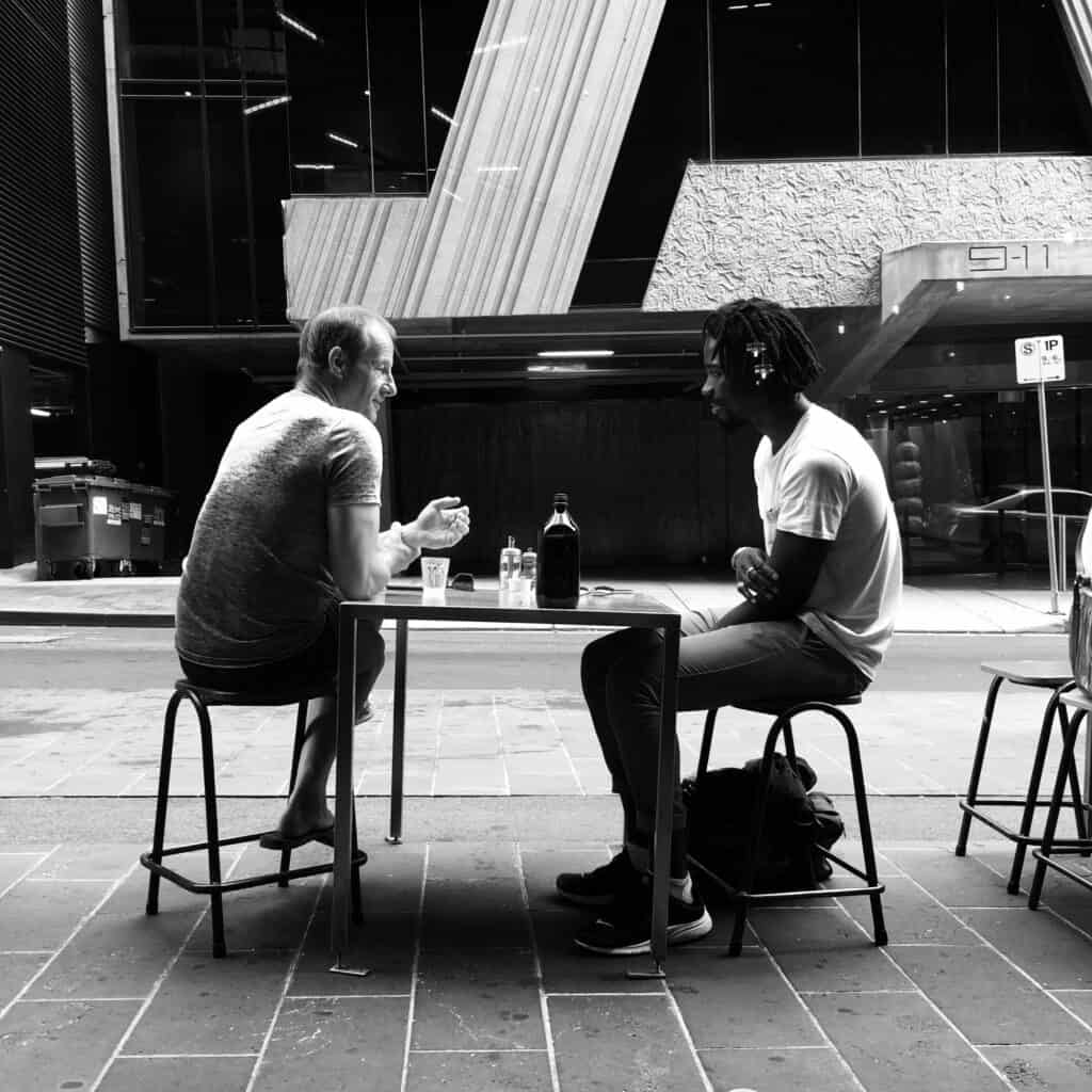 Two men sit at an outside table talking.