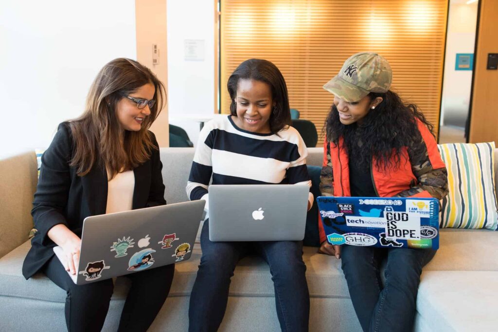 A group of three women sit with their laptops on their lap.