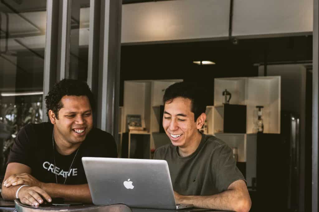 Two men sit in front of a Mac computer, laughing and smiling.