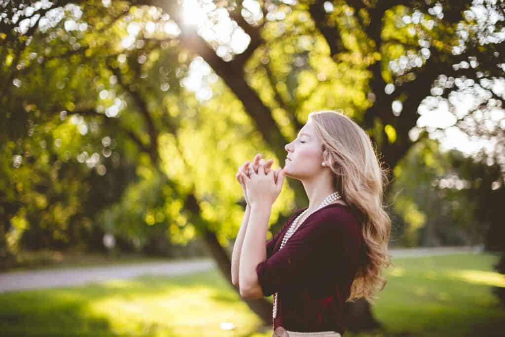 A white woman stands outside with her hands clasped together, eyes closed, and face toward the sun.