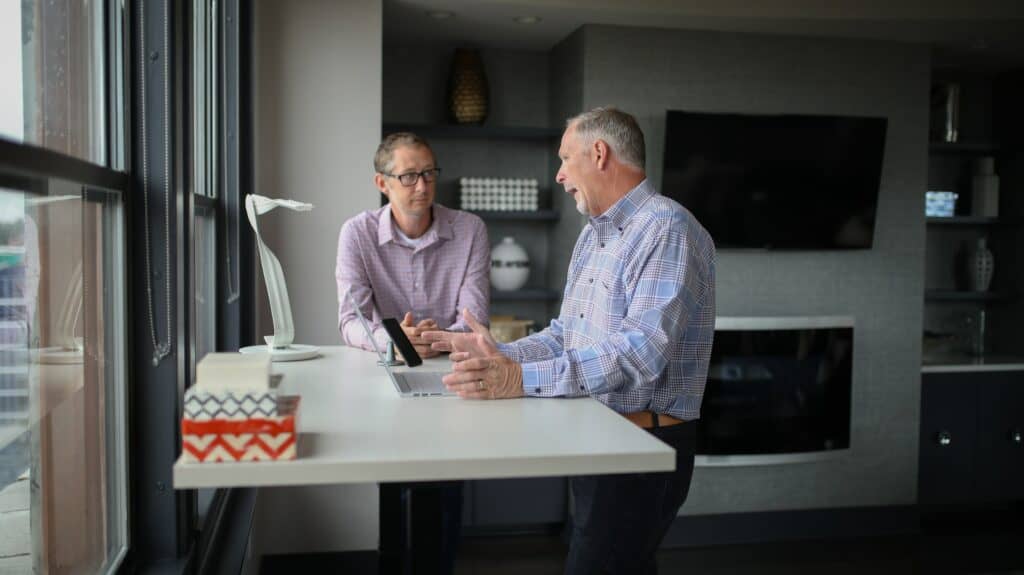 Two mature men stand at a work desk, talking
