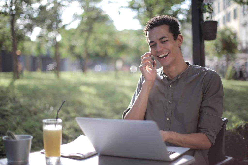 A man sits at a cafe with his laptop and orange juice as he talks on the phone