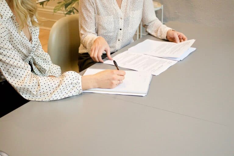 Two women sit at a table pointing to papers and signing them.