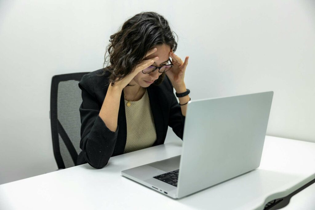 A woman sits in front of a laptop with her her hands on either side of her head.