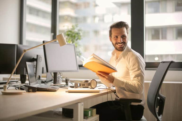 A man sits in front of a computer monitor, smiling, holding a book
