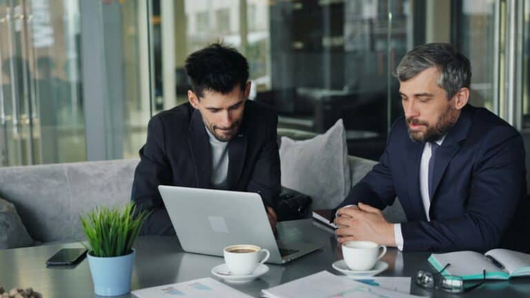 Two men sit outdoors at a table around a computer with coffee cups